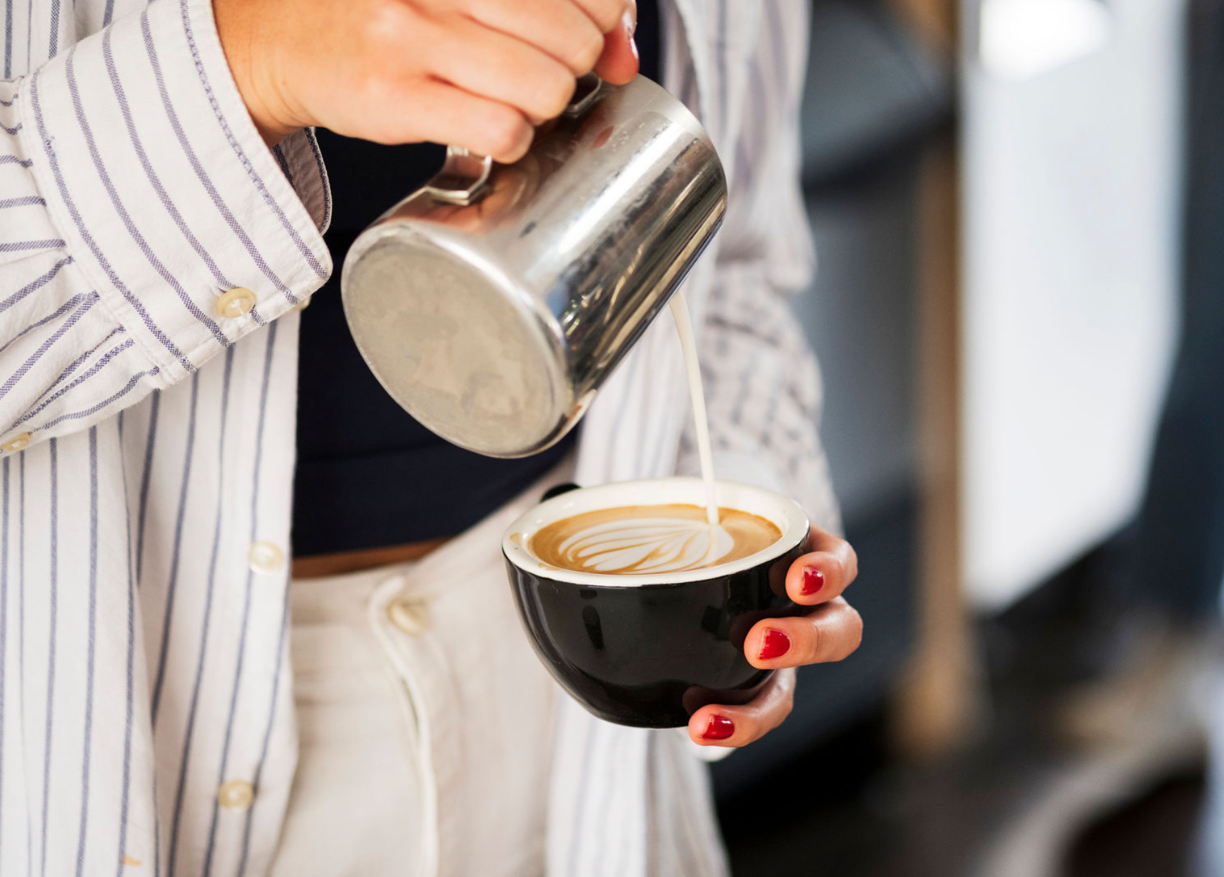 Person pouring steamed milk into a black coffee cup with a blurred background